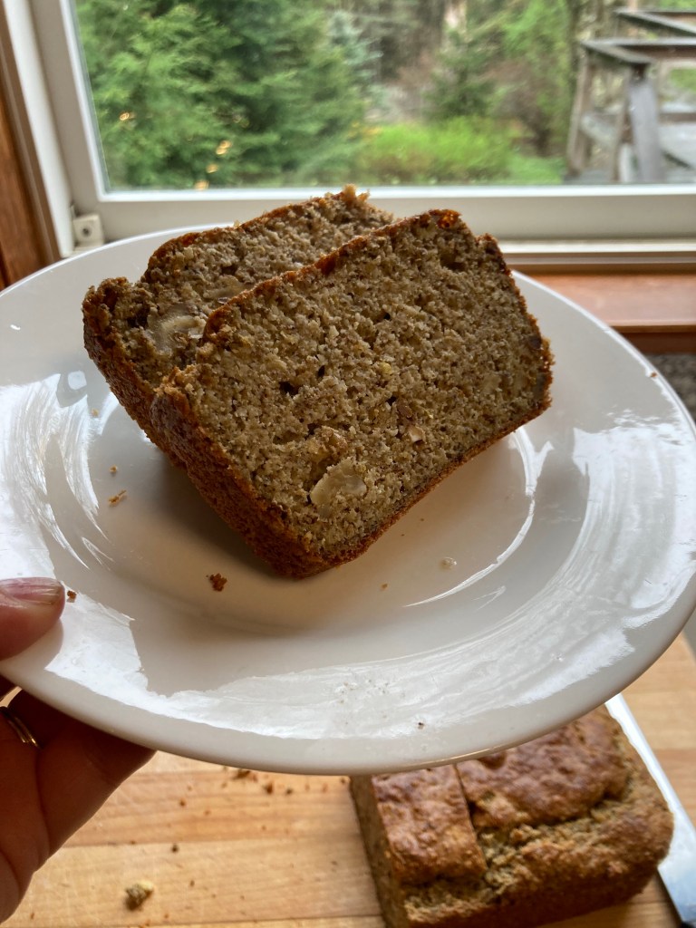 Two slices of chai-spiced protein bread with walnuts on a white plate, held by hand near a window overlooking a green forest